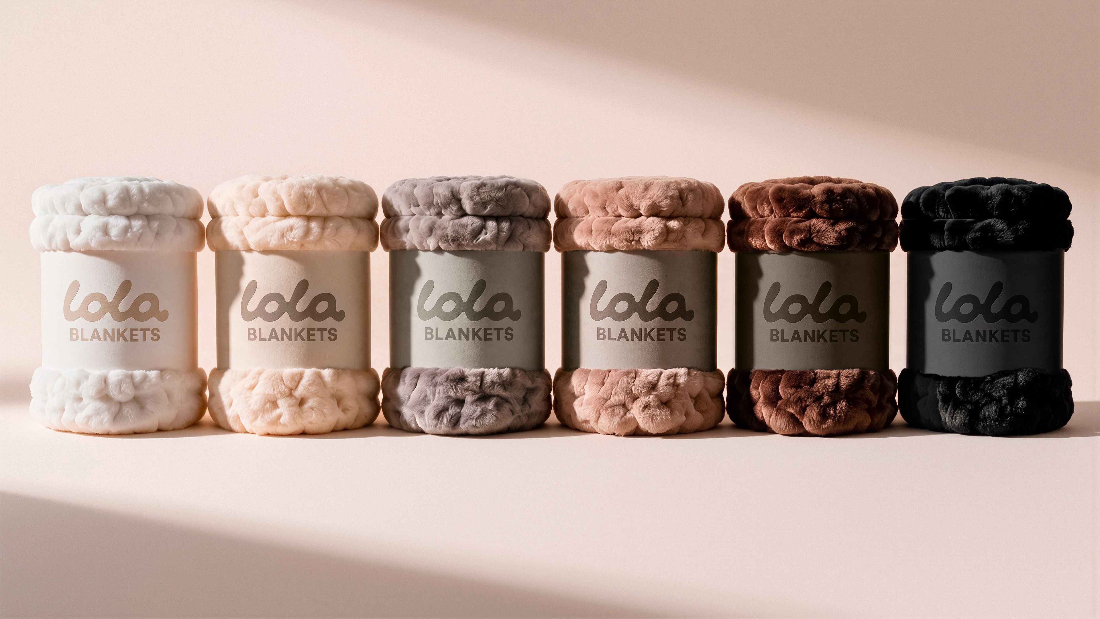Product packaging rendering for Lola Blankets featuring six rolled faux fur blankets in the full colorway lineup, each wrapped in an embossed Lola Blankets belly band, by TRUFFL branding agency.